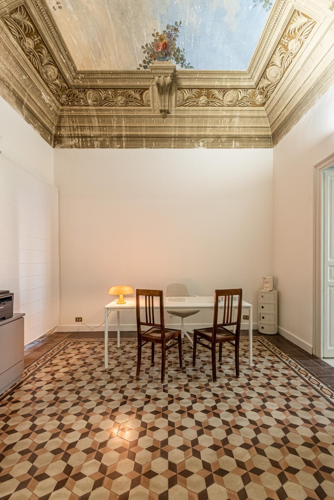 Stanza con soffitto decorato e pavimento decorato; al centro si trovano un tavolo e delle sedie, illuminati da una lampada.