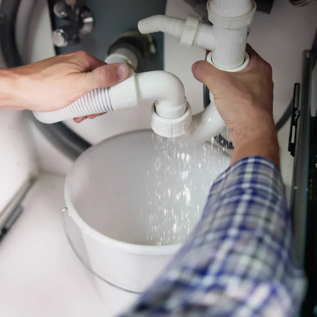 Hands fixing a white sink drain under a cabinet with water leaking into a bucket