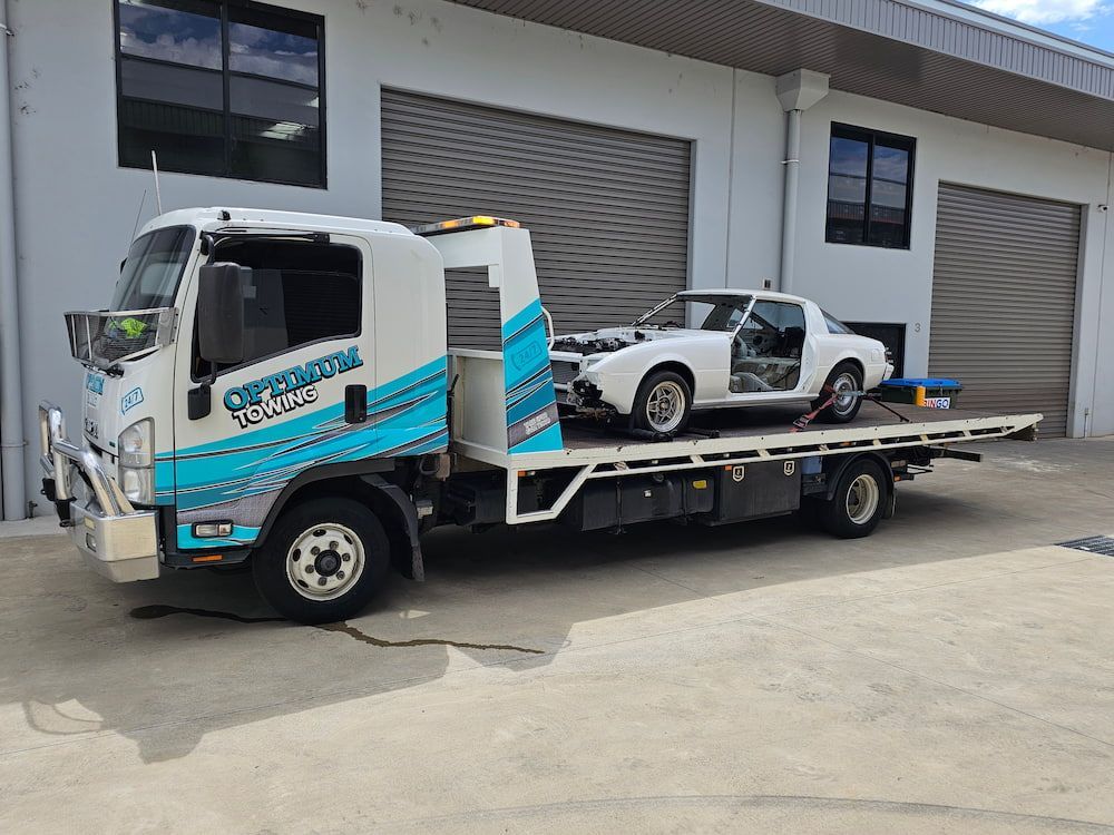 A Tow Truck With a White Car on the Back is Parked in Front of a Building — Optimum Towing in Gerringong, NSW