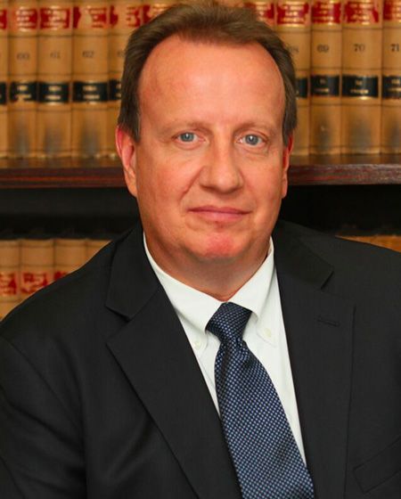 A man in a suit and tie stands in front of a bookshelf