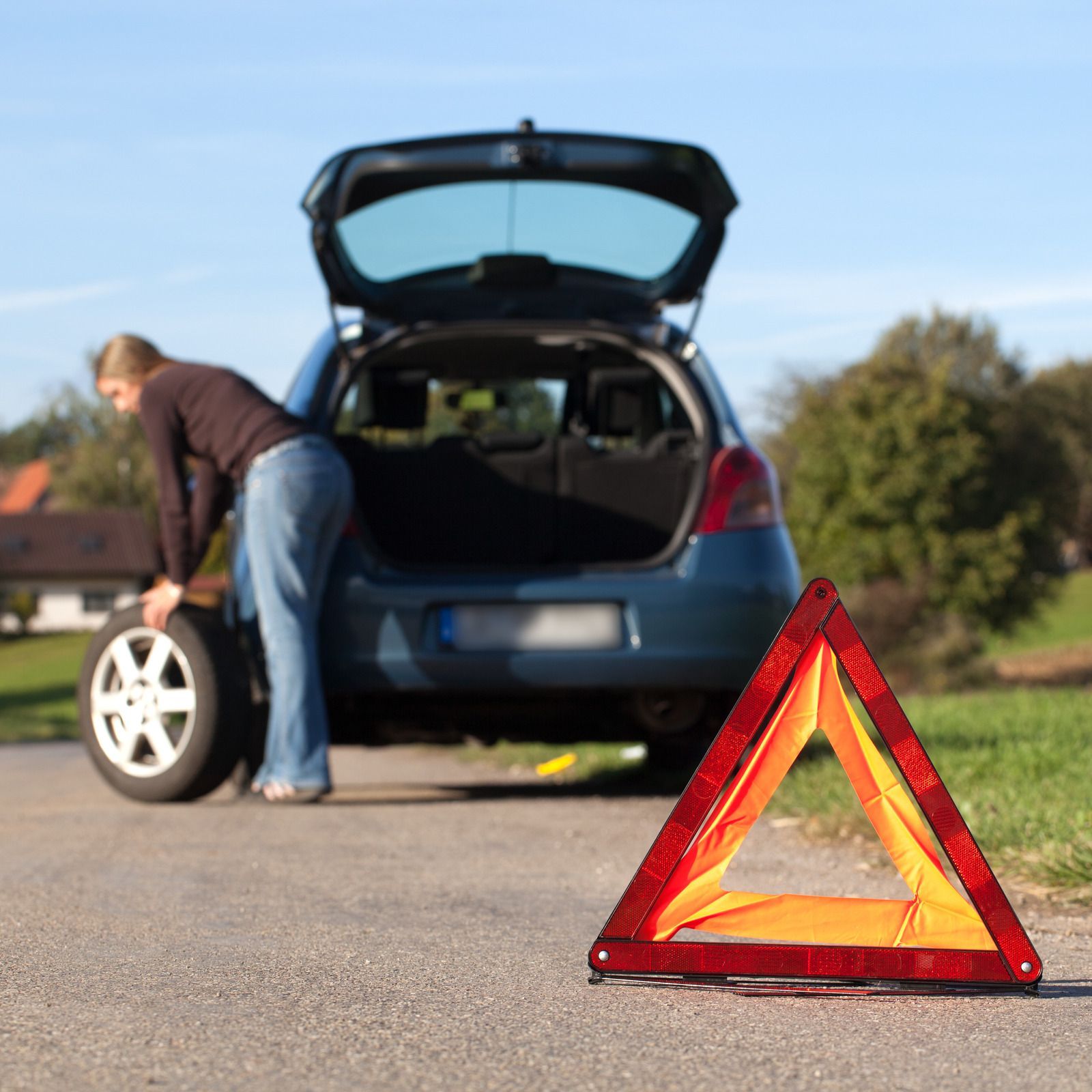 Woman changing a flat tire on a blue car on the side of a road, emergency triangle in foreground.
