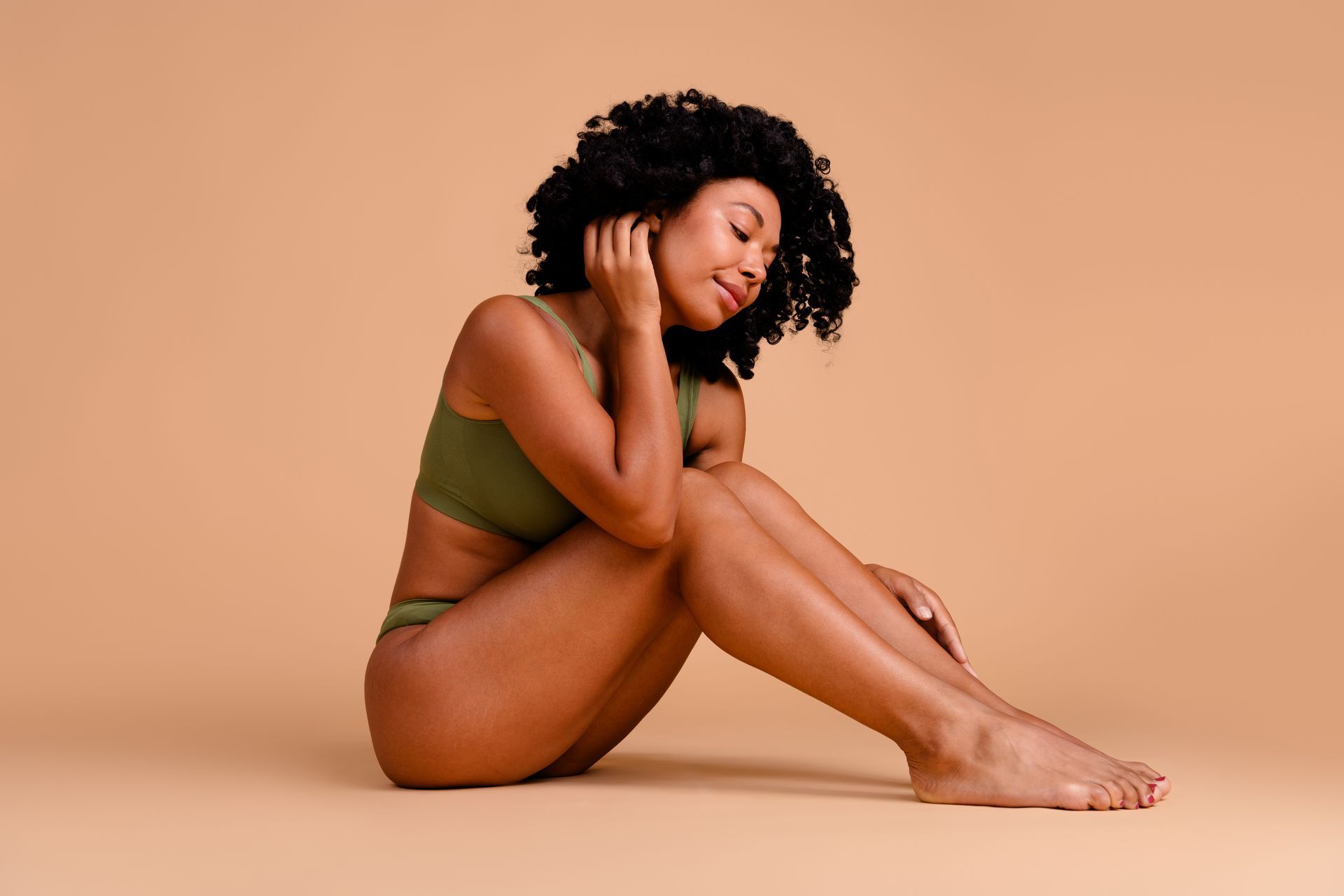 Woman in green bra and underwear, sitting with eyes closed, hands near face. Tan background.