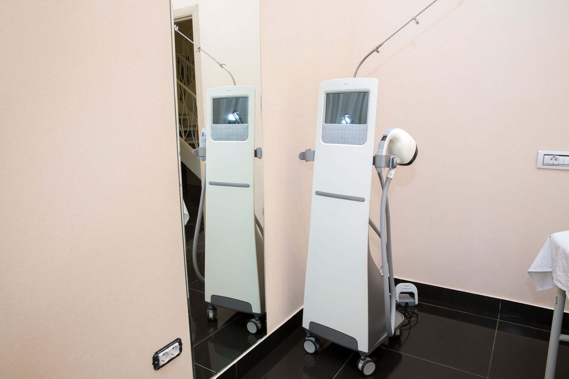 A tall medical machine with a screen stands by a mirror in a light-colored room.