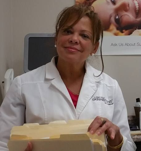 Woman in white coat holding files, smiling, in an office setting with a medical device in the background.