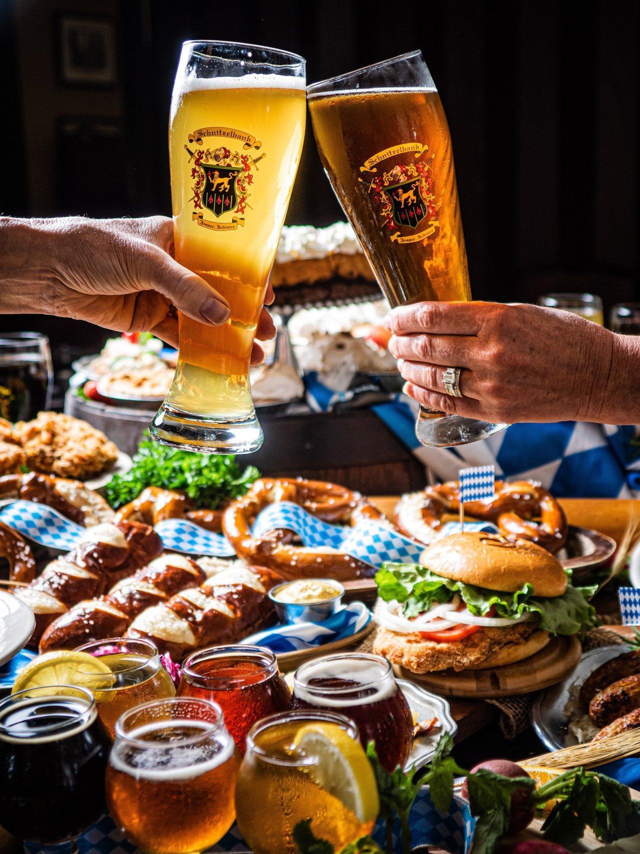 Hands clinking beer glasses over a table of pretzels, burgers, and dips in a cozy restaurant setting