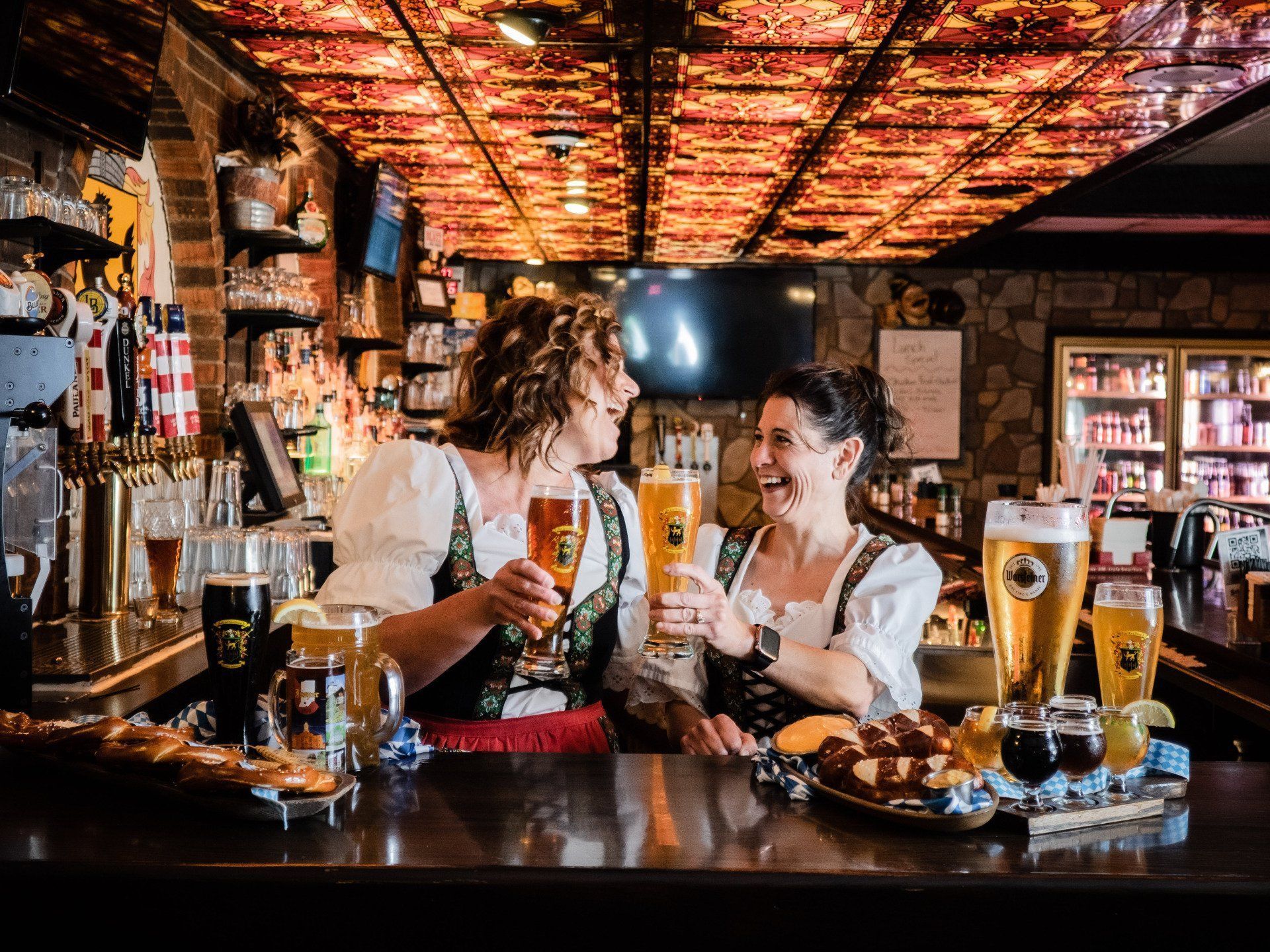 Two bartenders raise beer glasses behind a bar in a cozy pub with warm copper lighting
