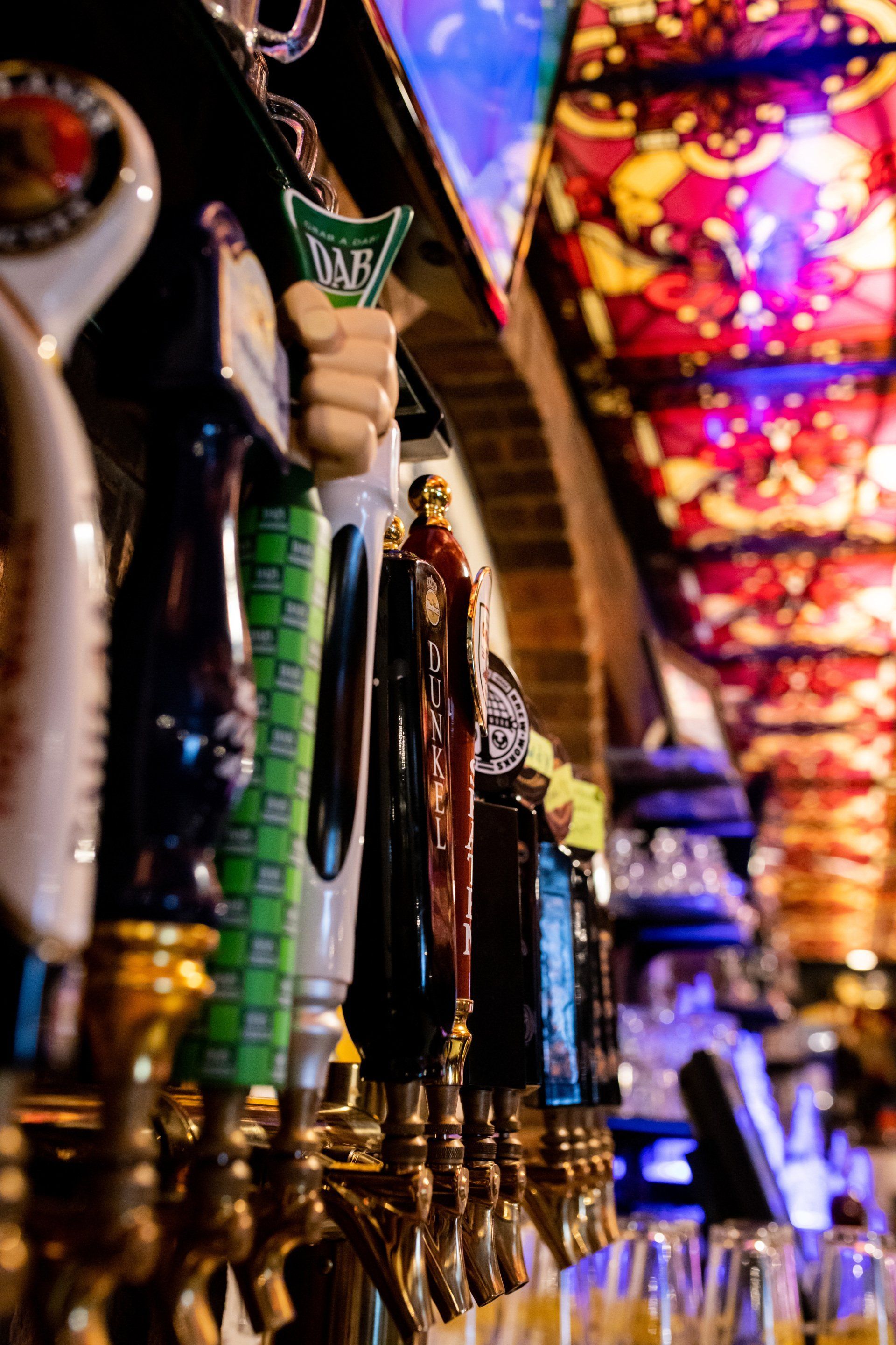 Row of colorful draft beer taps under a bright stained-glass bar ceiling