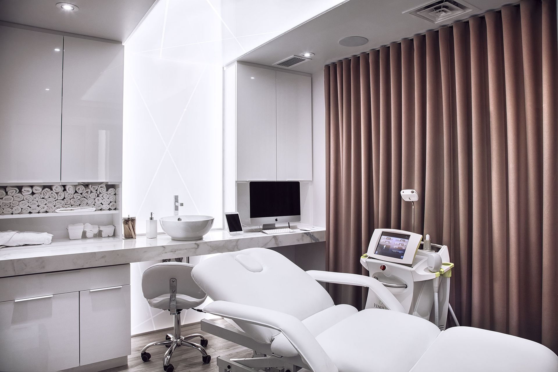 Une salle de spa médical lumineuse et moderne avec chaise de traitement, ordinateur et équipement cosmétique.