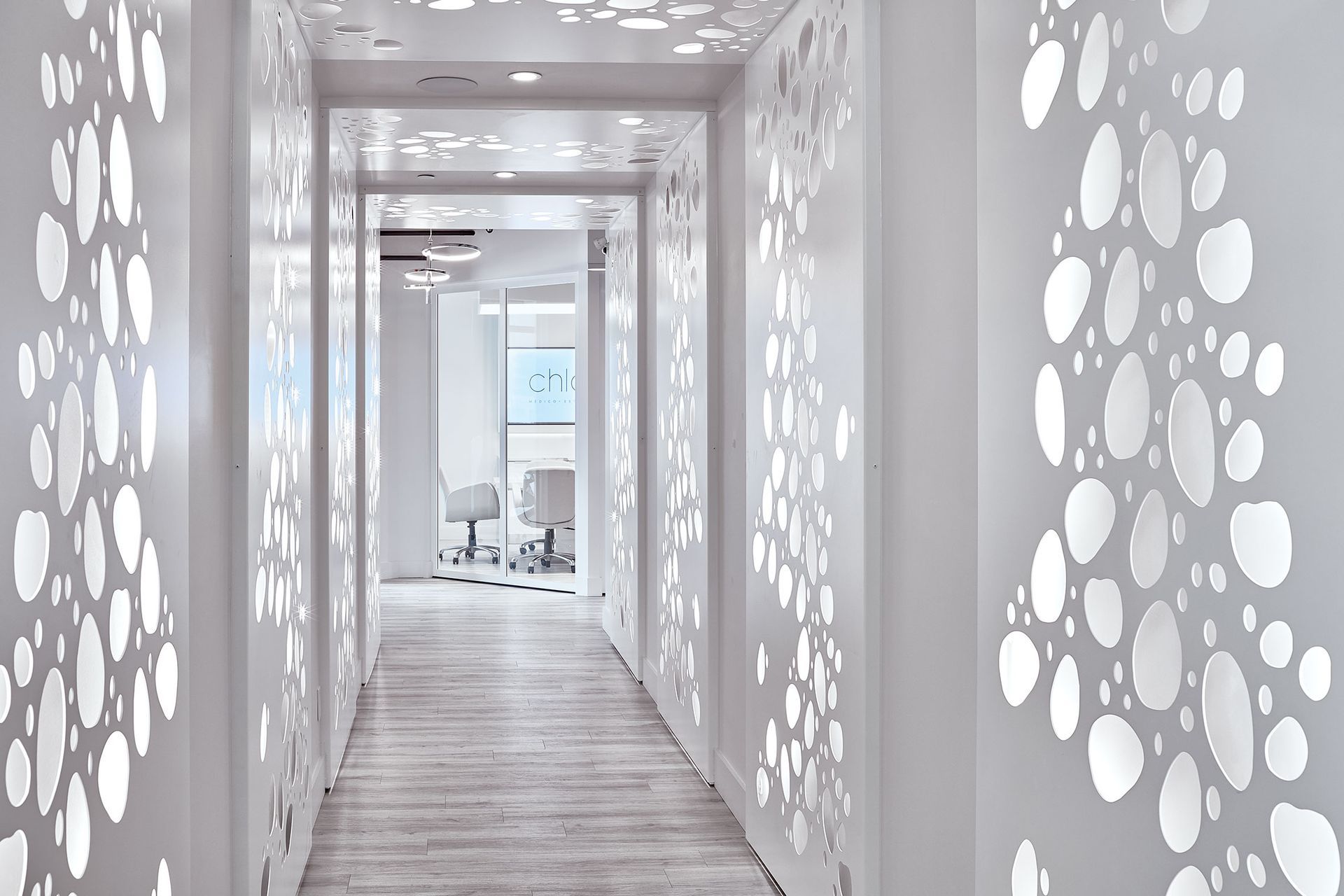 Un couloir blanc orné de découpes murales à motifs mène à une pièce lumineuse équipée de fauteuils dentaires.