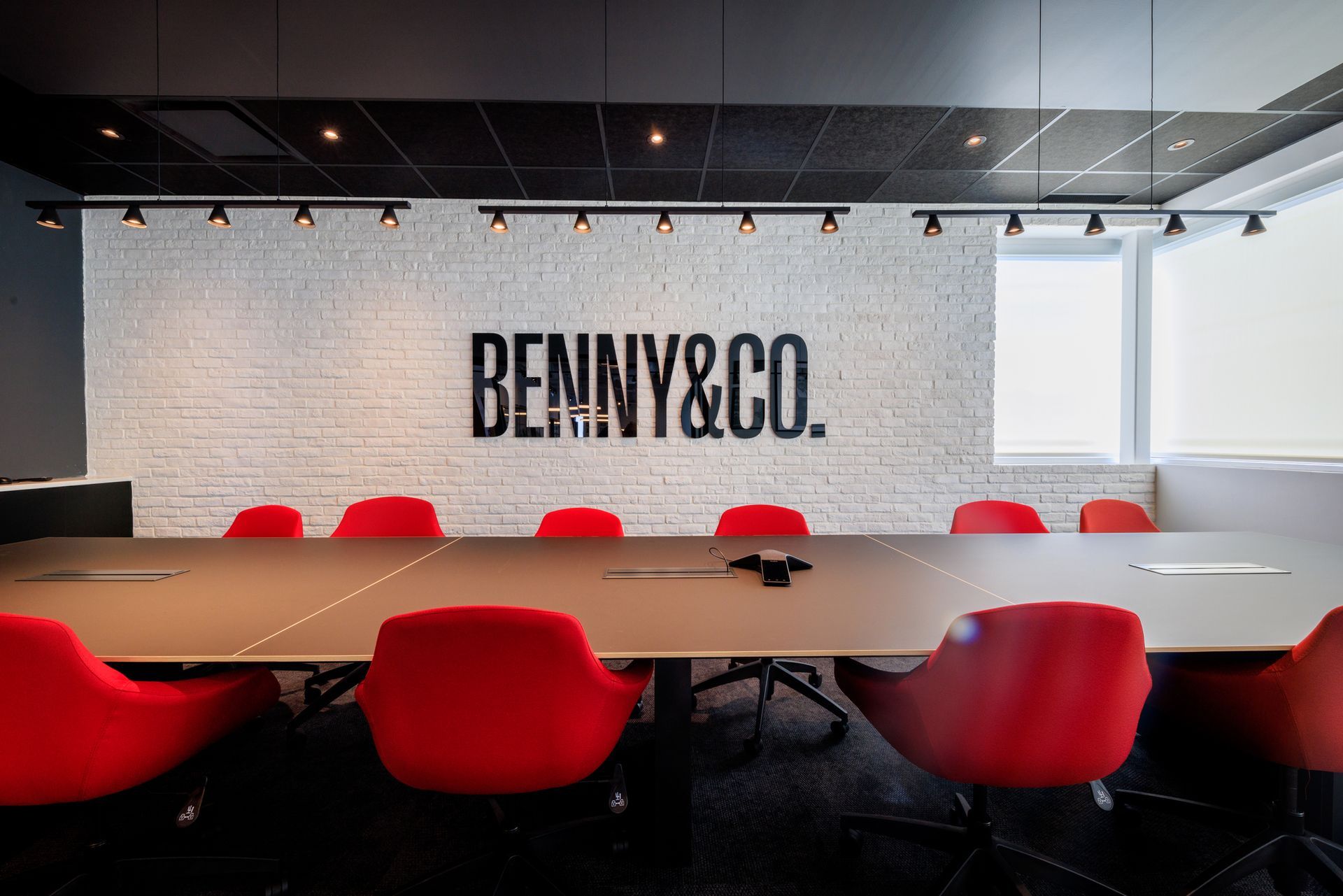 Salle de réunion avec longue table, chaises rouges, mur de briques blanches et logo