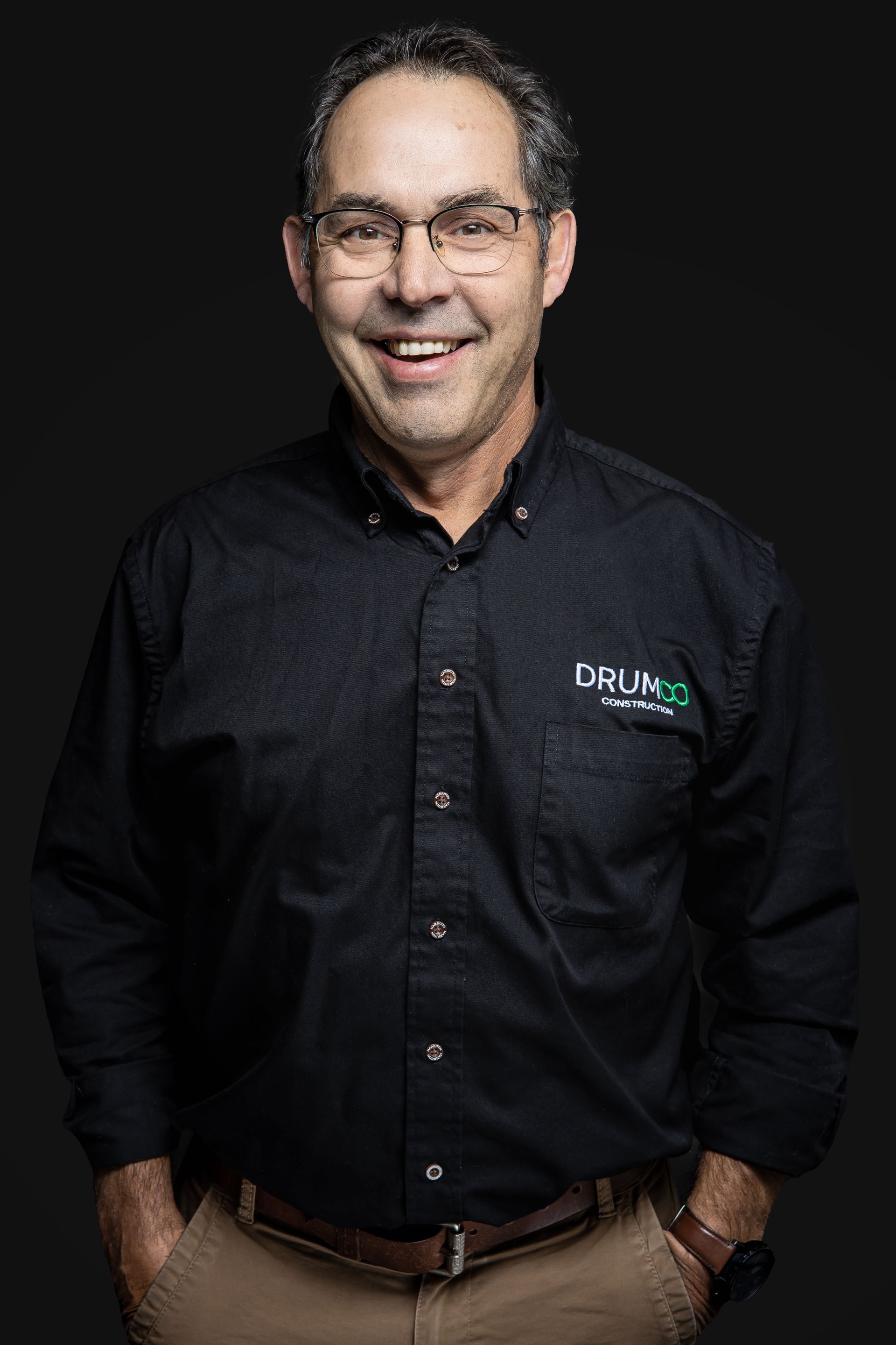 Un homme portant des lunettes et une chemise noire sourit avec ses mains dans ses poches.