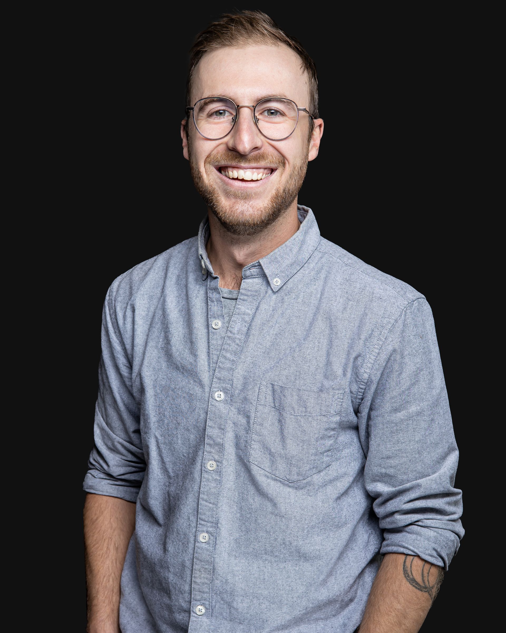 Un homme portant des lunettes et une chemise bleue sourit à la caméra.