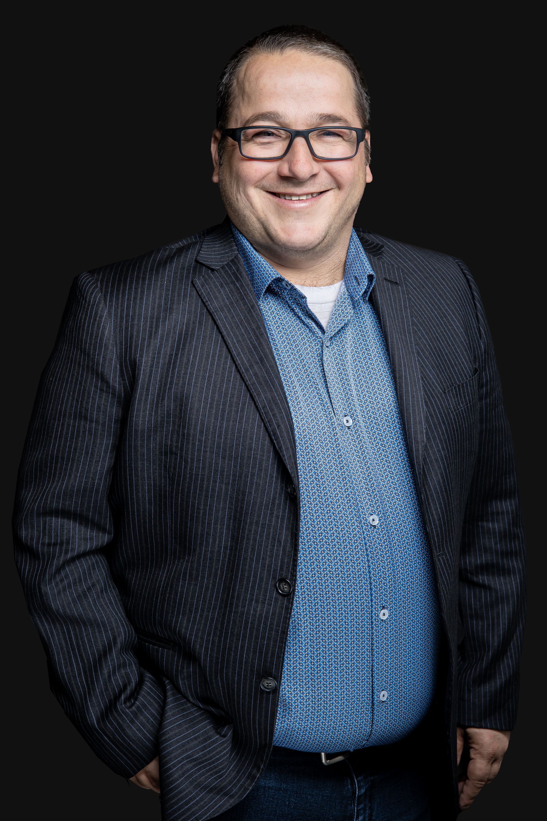 Un homme portant des lunettes et une veste de costume sourit à la caméra.