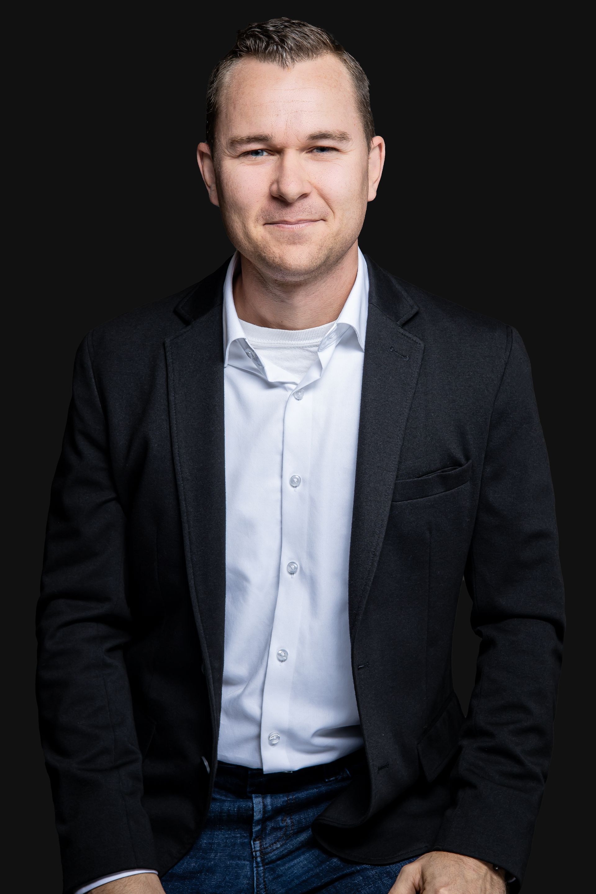 Un homme en costume et chemise blanche est assis devant un fond noir.