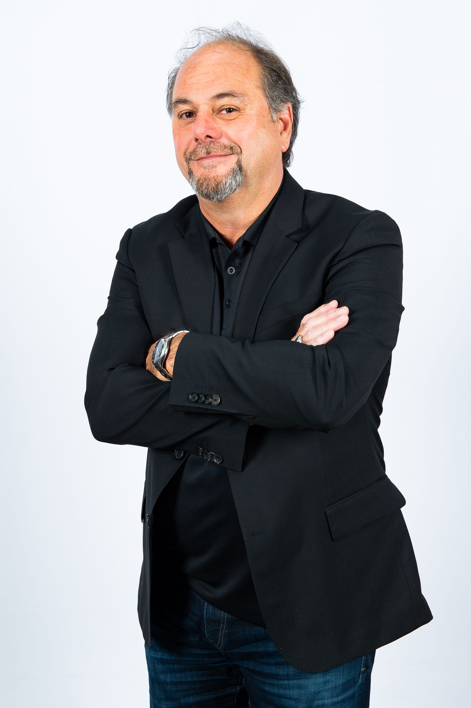Un homme portant une veste noire et un jean est debout, les bras croisés.