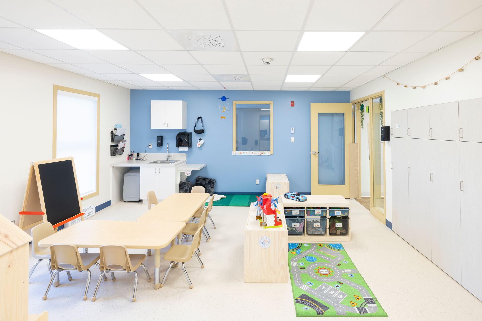 Salle de classe lumineuse avec table et chaises, mur d'accent bleu, rangements et aire de jeux.