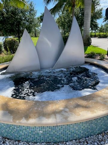 Fountain with three silver triangular structures, water splashing, surrounded by a mosaic rim and landscaping, outdoors.