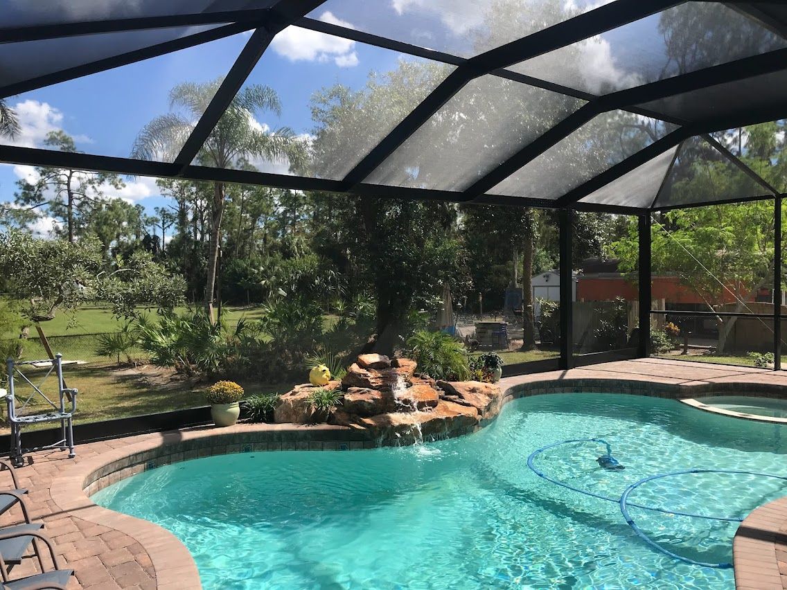 Swimming pool with waterfall feature, under a screened enclosure, with a view of trees and sky.