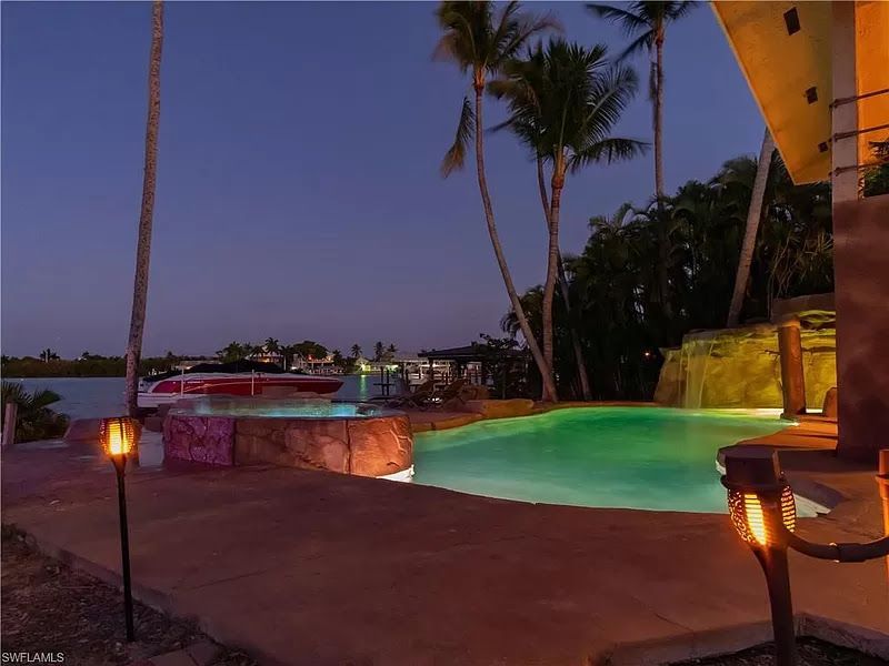 Pool and waterfront view at dusk. Palm trees, boat, and tiki torches.