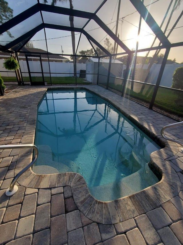A rectangular swimming pool surrounded by brick, under a screened enclosure. Bright sunlight overhead.