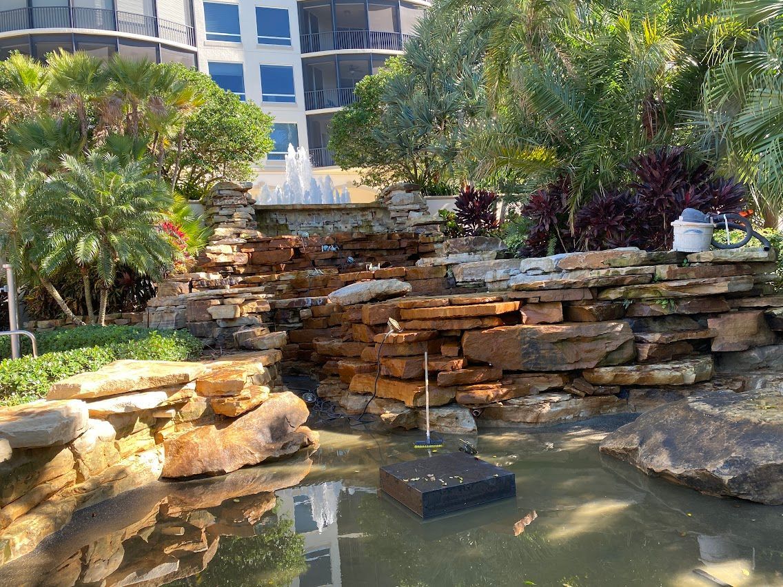 Waterfall feature in a landscaped area with a pond. Stone tiers, green plants, and a building in the background.