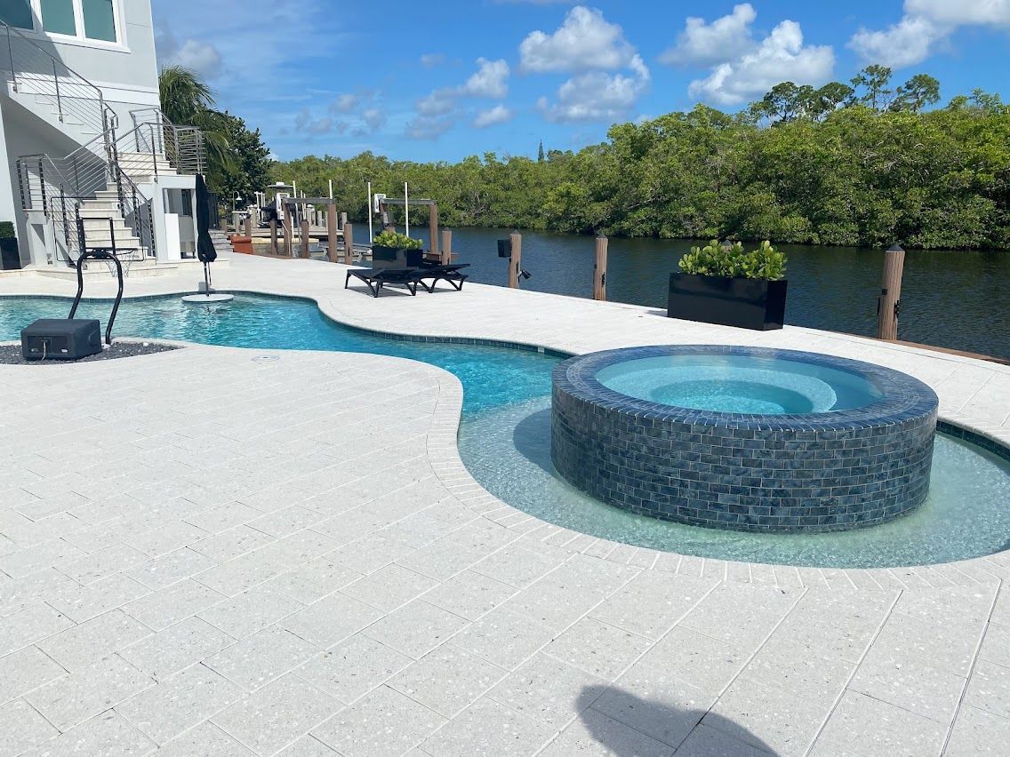 Pool and hot tub area with blue water, surrounded by white stone patio, and trees in the background.