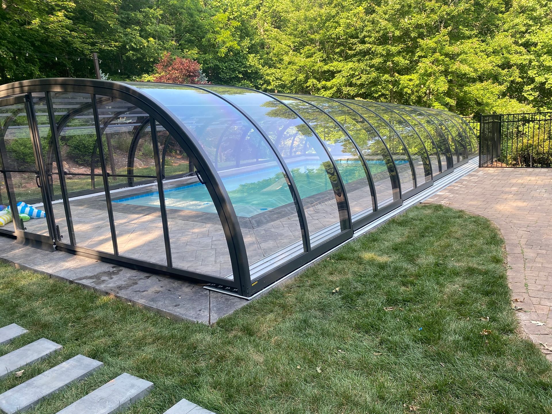 A pool covered by a transparent enclosure, in a backyard setting with green grass and trees.