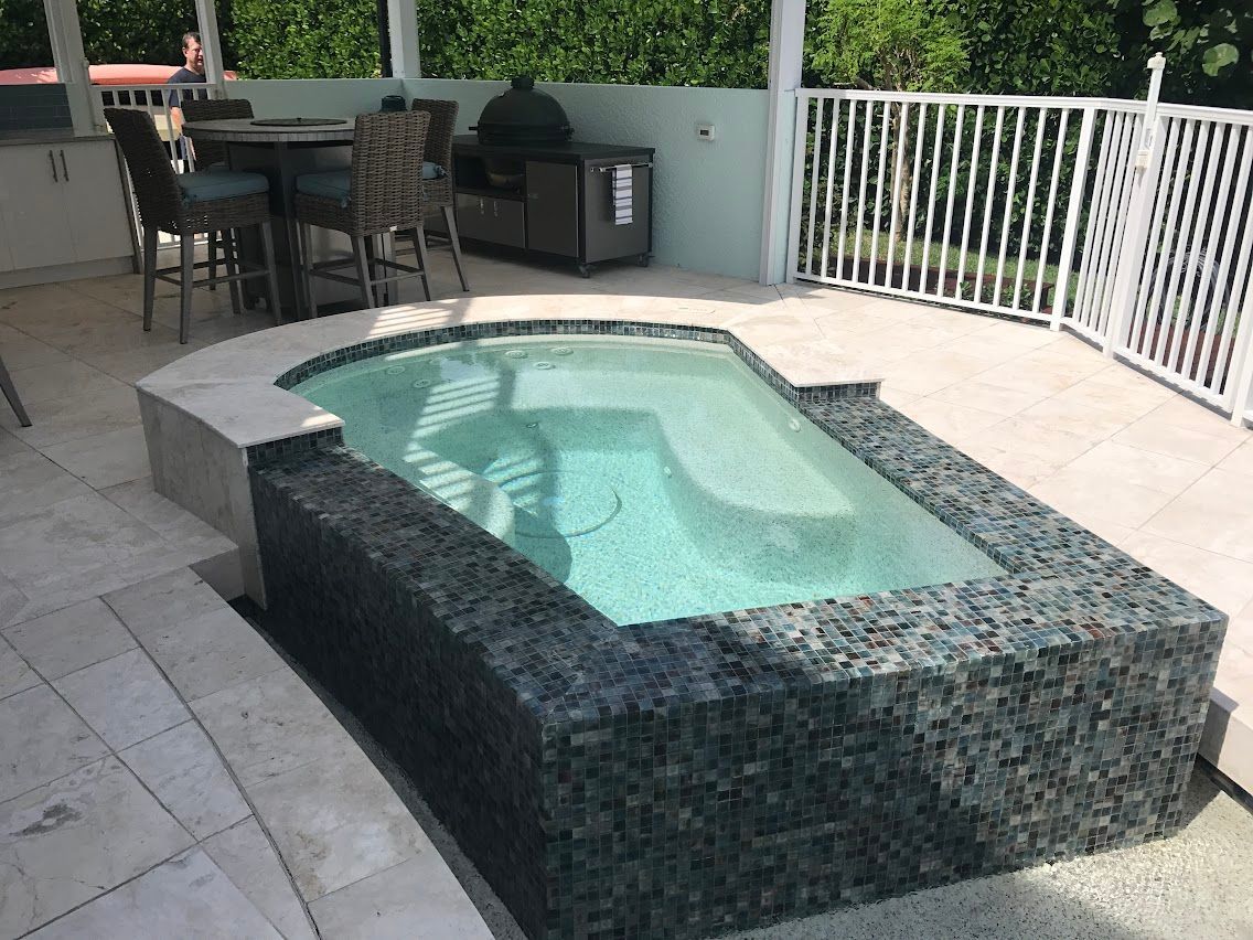 Hot tub surrounded by stone and mosaic tile on a patio, with a dining set and railing visible.