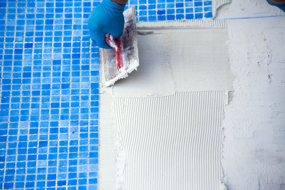 Person in blue glove using a notched trowel to apply white adhesive on a surface for installing blue mosaic tiles.
