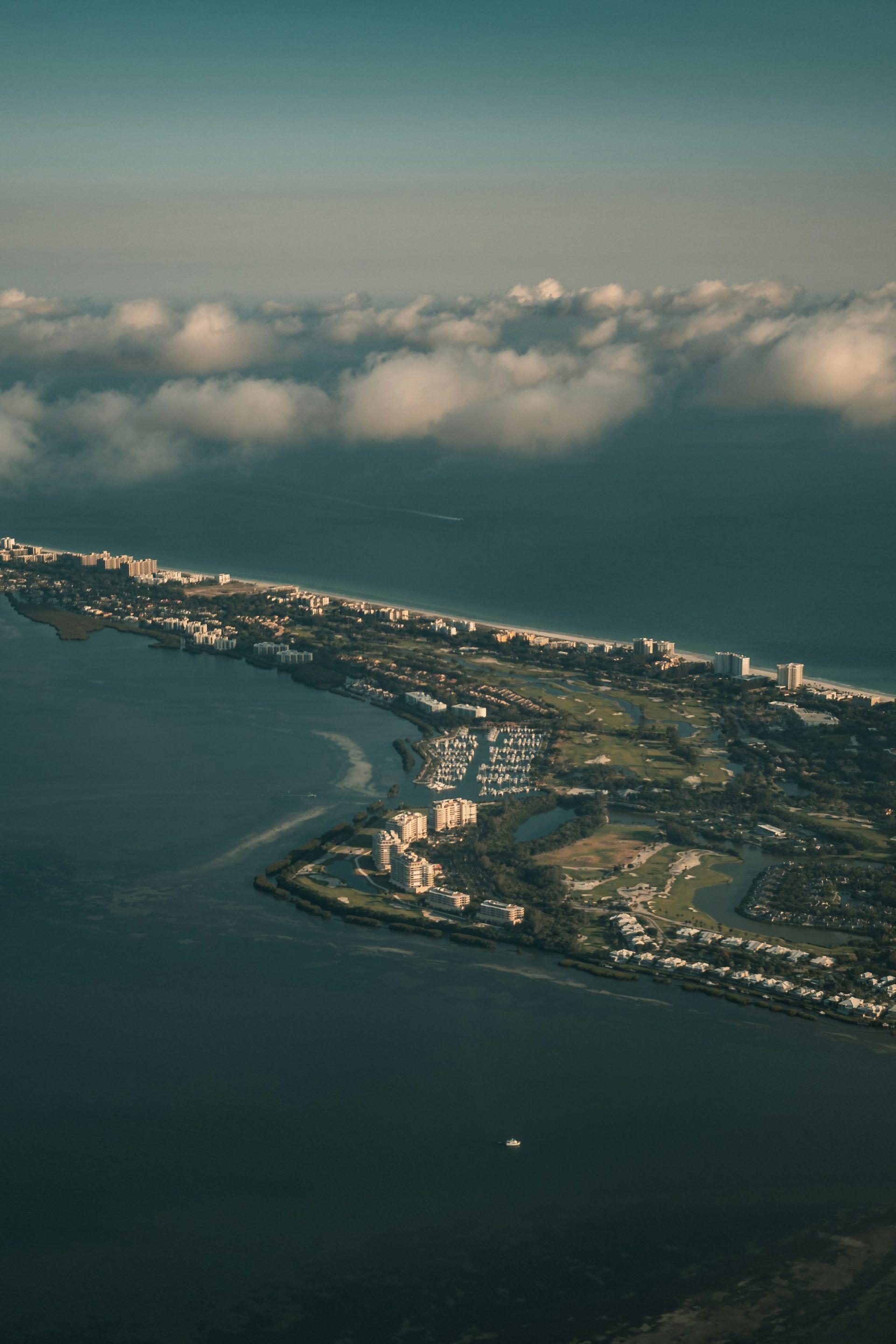Aerial view of a coastal strip with buildings, trees, and beaches, surrounded by water and partially covered by clouds.