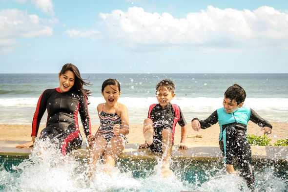 A woman and three children splashing in pool with ocean in background. They are laughing and wearing swimsuits, enjoying a sunny day.