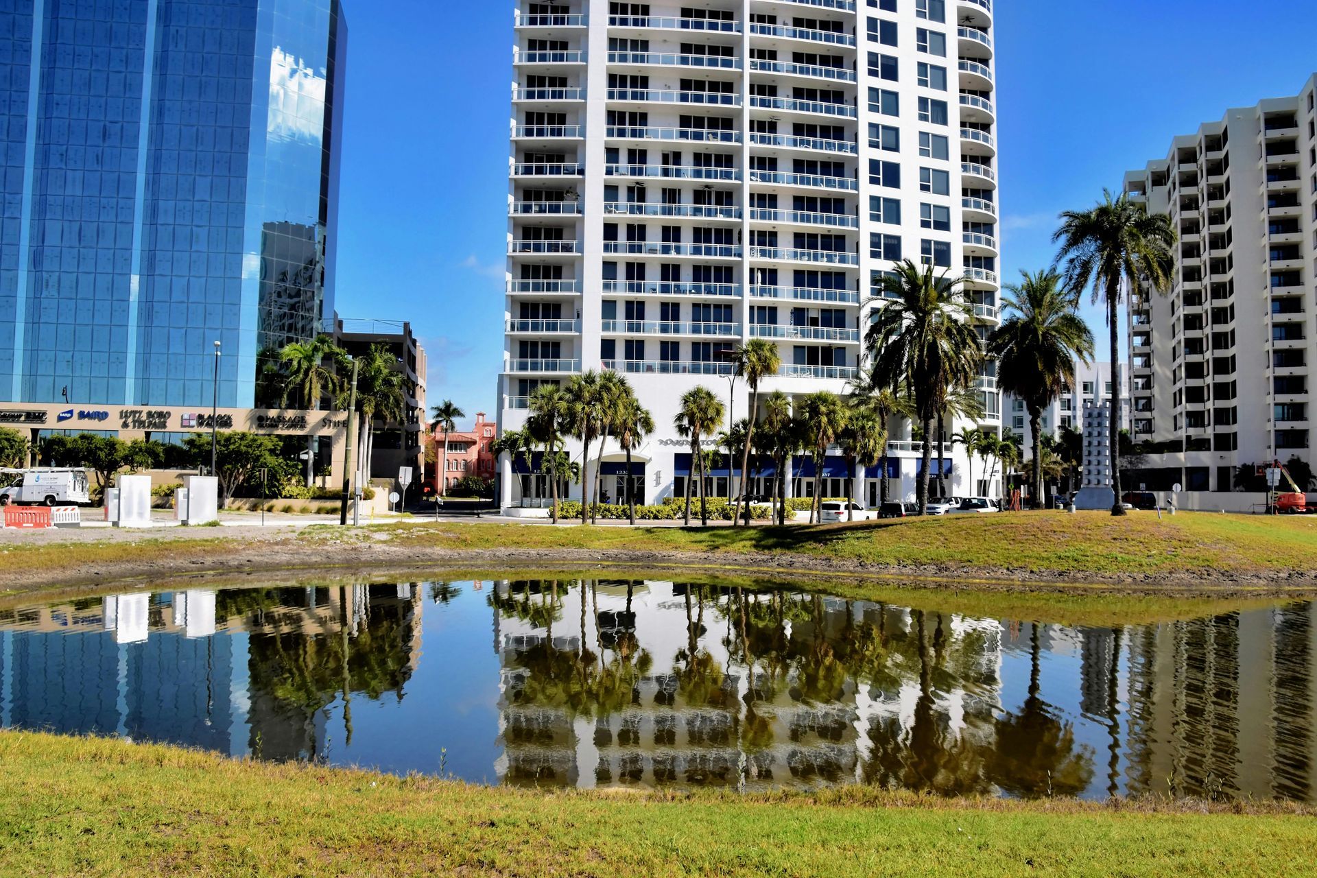 Buildings reflected in a calm body of water; palm trees and green grass in the foreground, under a blue sky.