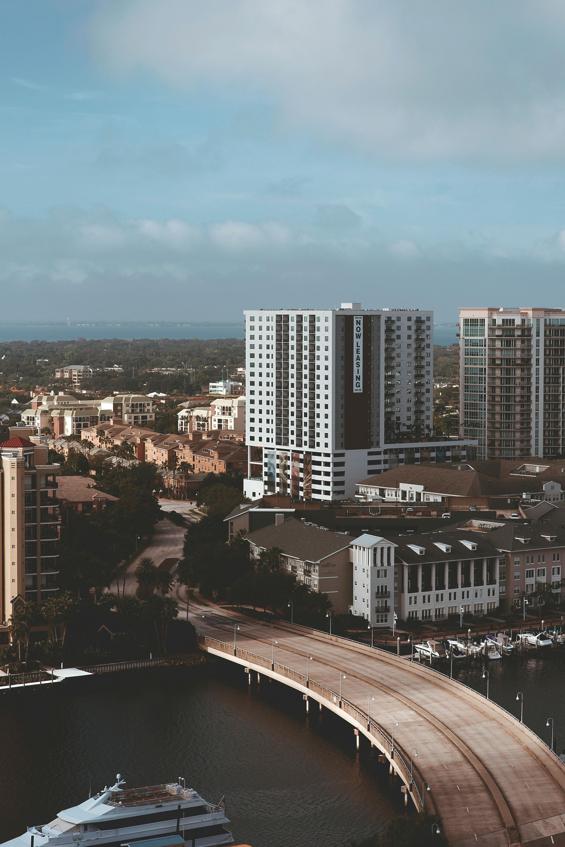 Cityscape with a bridge over water, featuring tall buildings and a cloudy blue sky.