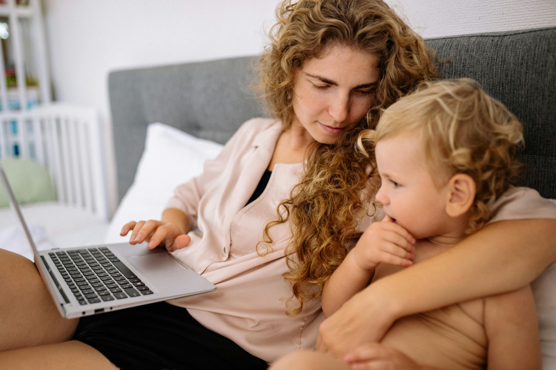 Woman and toddler on a bed, the woman working on a laptop. Woman is focused, and the child looks curious.