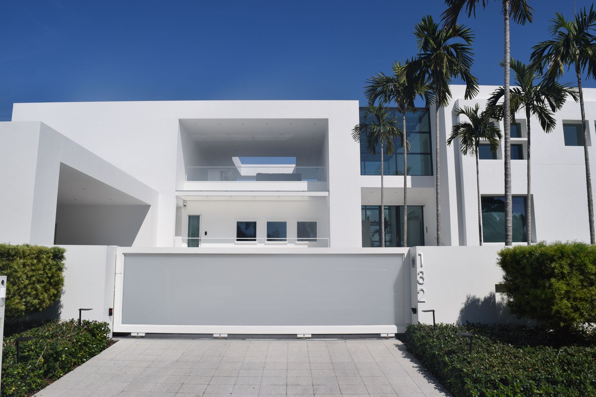 Modern white house with a large grey sliding gate and palm trees against a blue sky.