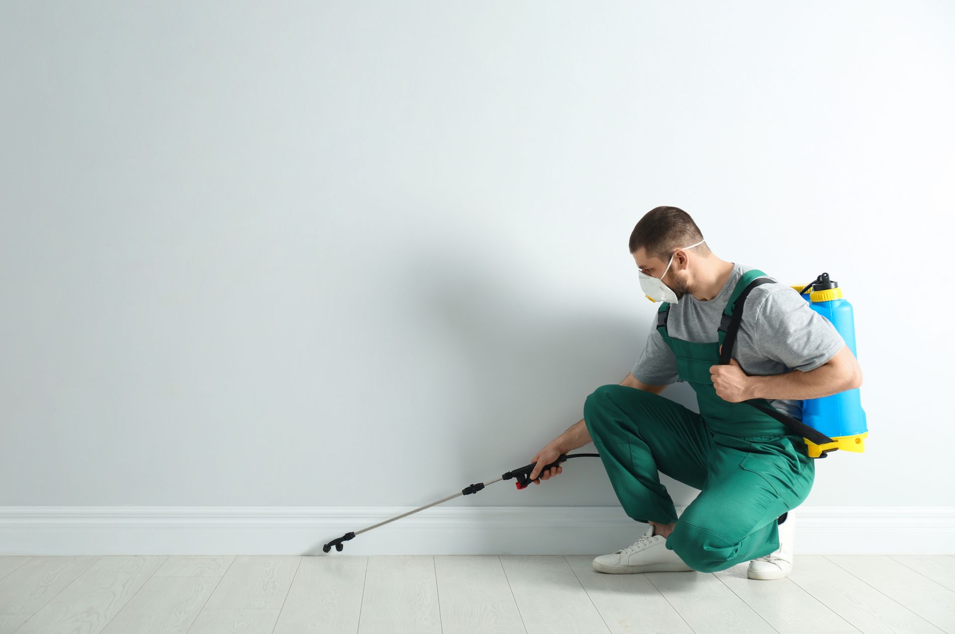 A pest control worker in uniform spraying insecticide along a wall baseboard