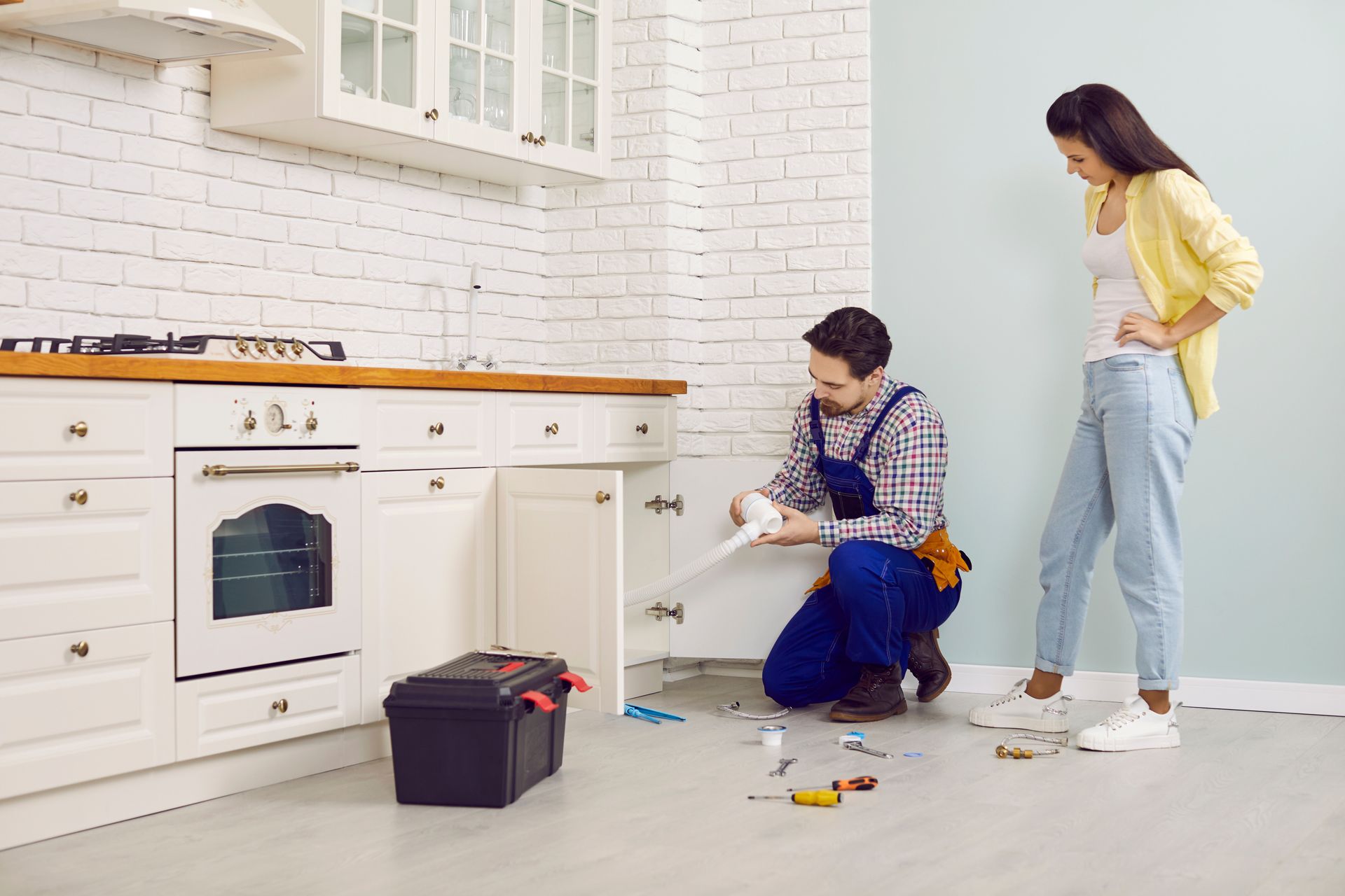 Plumber fixing some problems with the sink drain. Young lady calls the plumber to help her unclog the clogged sink pipe in the modern white kitchen of her house Plumber fixing some problems with the sink drain.