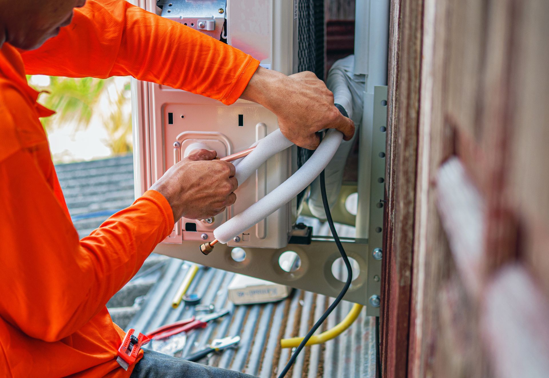 Technician installing AC unit, connecting pipes and cables on exterior wall of building.