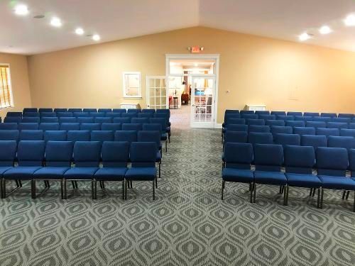 Rows of blue chairs in a beige-walled room, arranged for seating, with a doorway visible at the back.