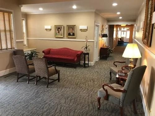 Hallway with seating, red sofa, portraits, and a lamp. Light-colored walls and carpet.