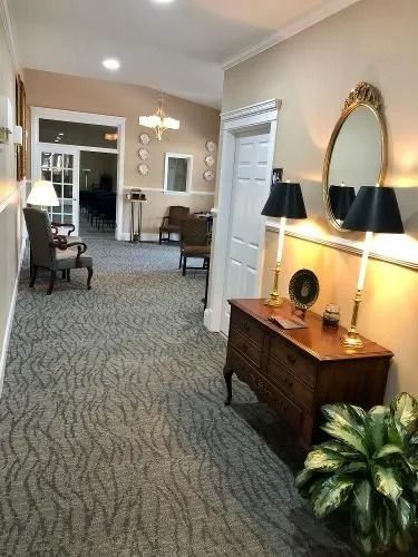 Hallway with furniture, mirror, lamps, and a view of a room with chairs. Beige walls, patterned carpet.