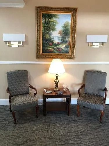 Two chairs face each other with a small table and lamp between them under a painting and two wall lights.