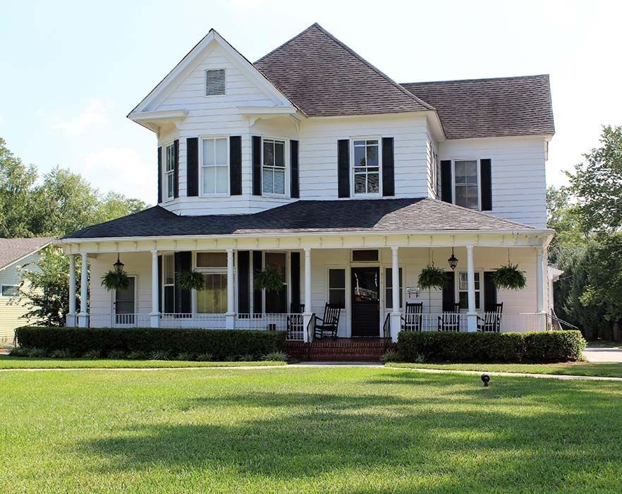 White two-story house with a wraparound porch, black shutters, and a well-kept lawn under a sunny sky.