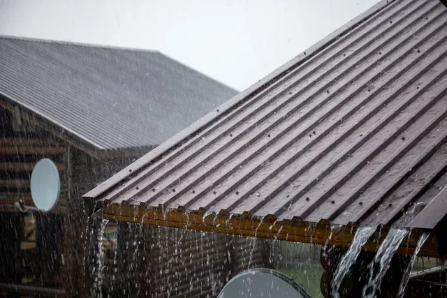 Roofer on a metal roof, hammering a piece of flashing. He wears a hard hat and work clothes.