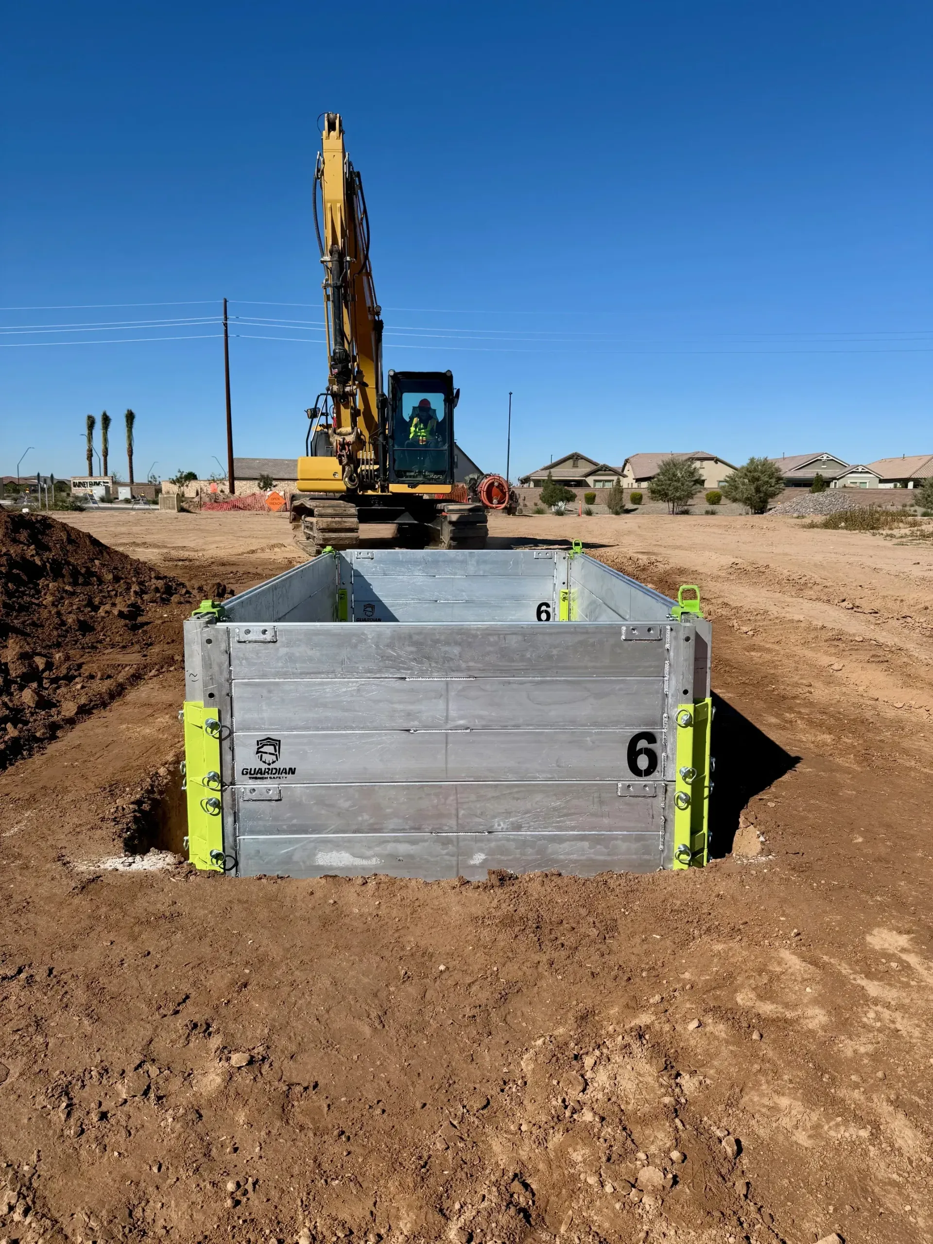A yellow excavator parked behind a rectangular metal trench shield box in a dirt lot under a clear blue sky.