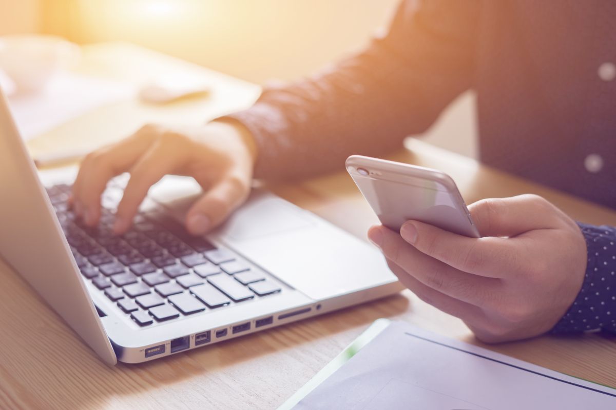 A person is typing on a laptop while holding a cell phone.