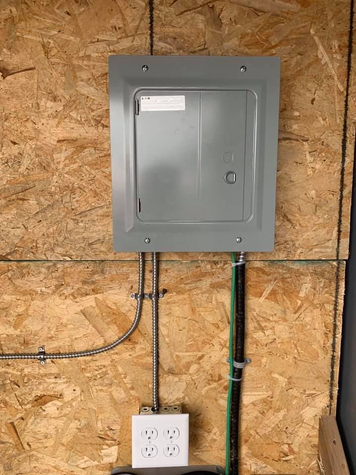 A electrical box is hanging on a wooden wall next to two electrical outlets.