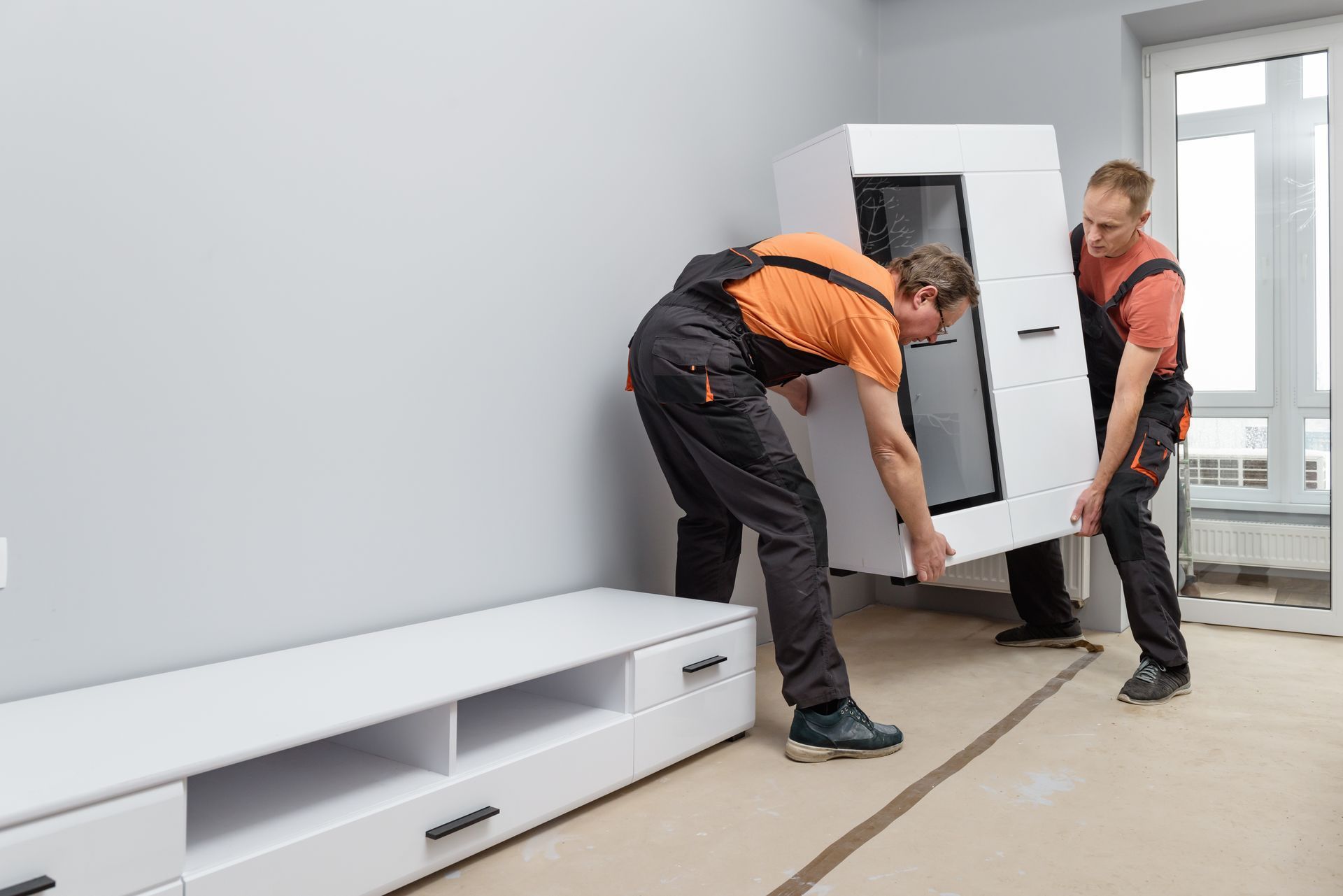 Two men are carrying a white cabinet in a living room.