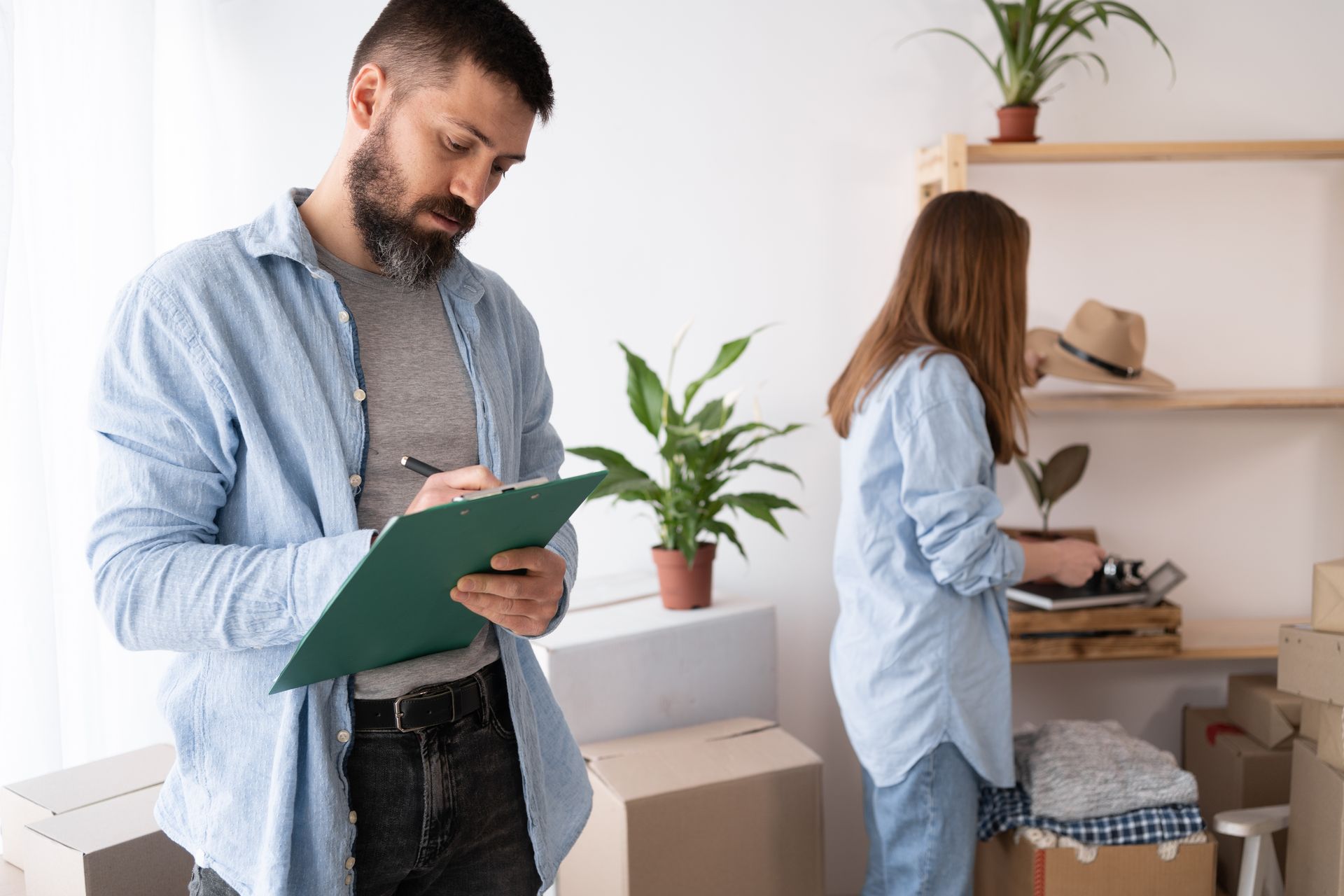 A man is holding a clipboard while a woman is packing boxes in a room.