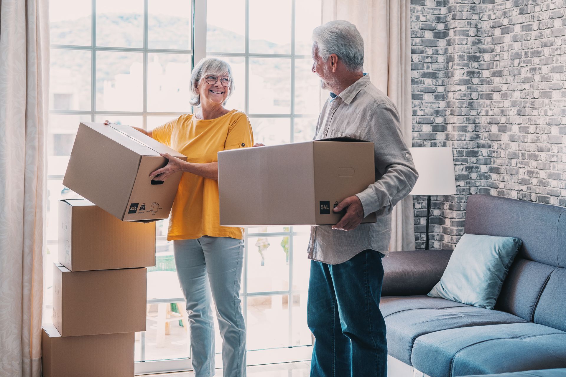 Elderly couple carrying moving boxes in a brightly lit living room, smiling.
